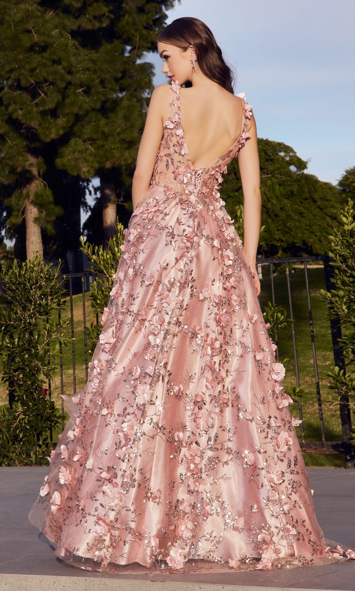 A woman stands outdoors in the Long Glitter-Floral A-Line Prom Ball Gown J838, a floor-length pink dress with floral glitter appliqué, deep V-back, and flared skirt. Trees and greenery fill the background.